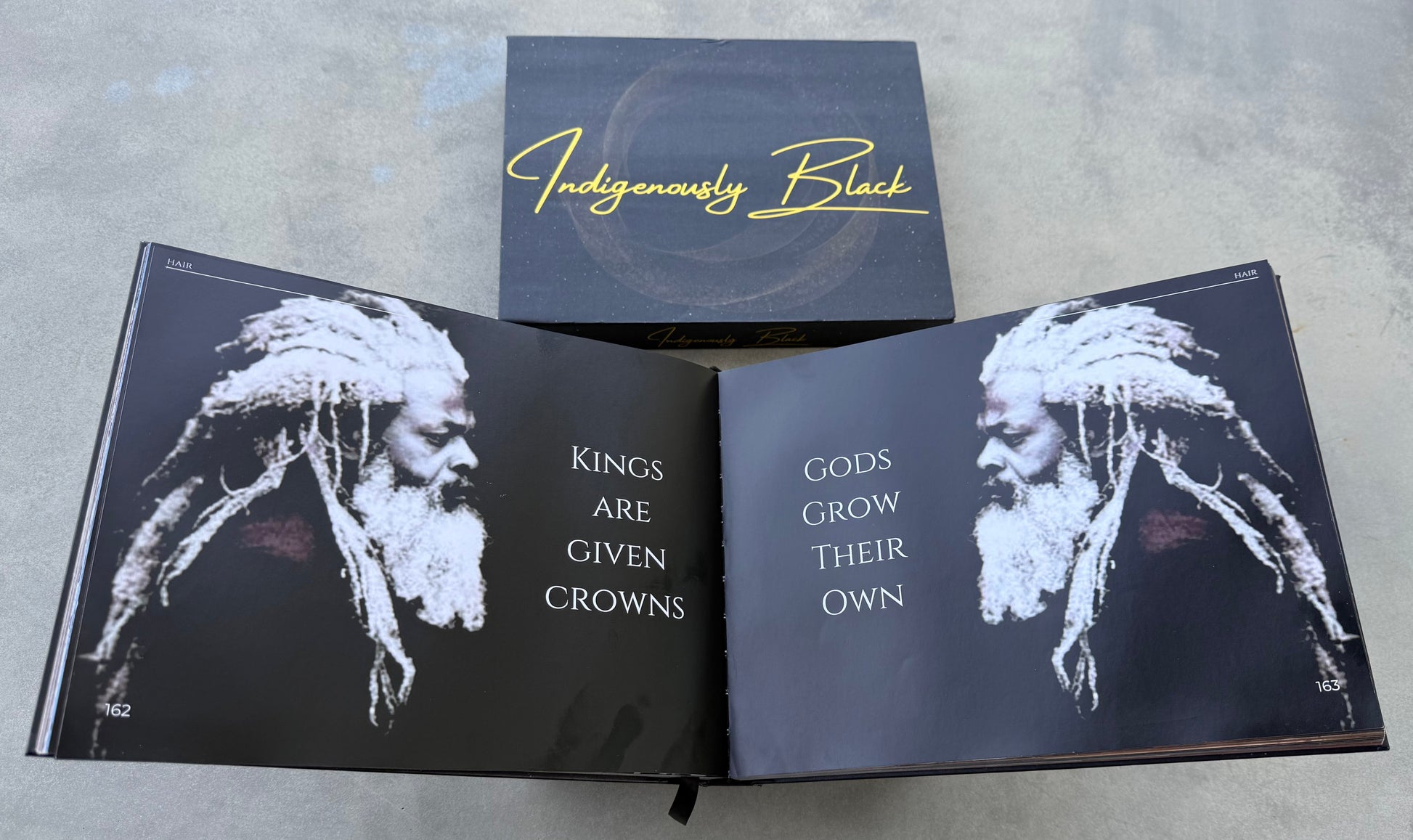 Open book with black and white illustrations and text on a gray surface, featuring the brand 'Indigenously Black'.